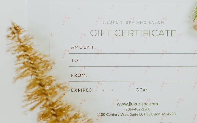 A J. Jukuri gift certificate laid on a pale surface beside a dried flower stem